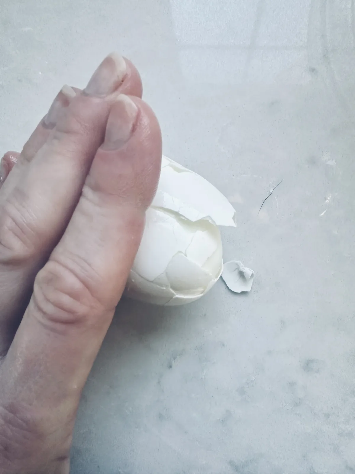A hand cracks a hard-boiled egg on a light-colored surface, with pieces of eggshell breaking off and scattered nearby.