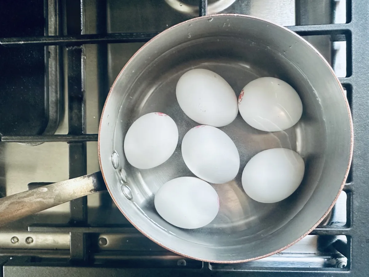 Six eggs are boiling in a pot of water on a stove, with the pot partially filled and the eggs submerged. The stove grates and control knobs are visible.