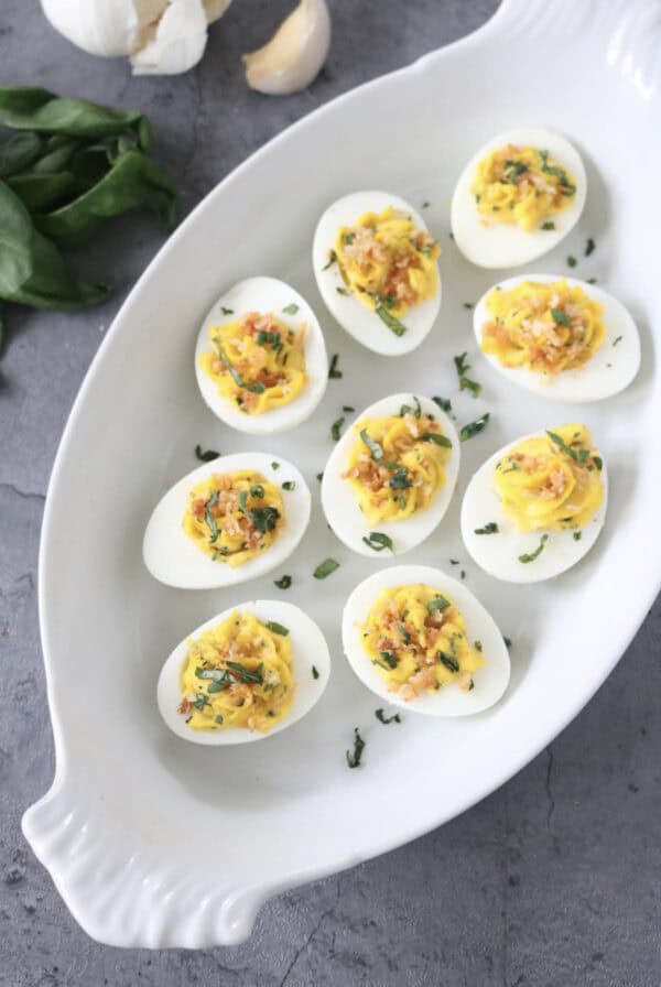 A white oval dish with ten deviled eggs, topped with herbs and spices, sits on a gray textured surface. Garlic cloves and fresh green leaves are nearby.