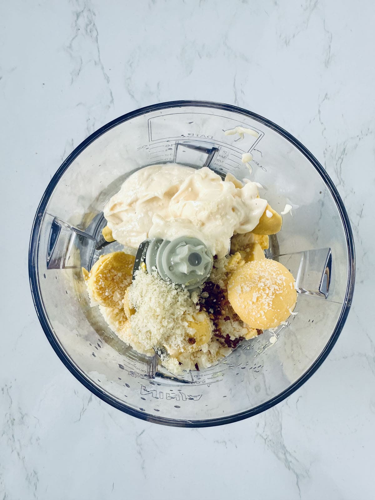 A top-down view of a blender filled with mayonnaise, egg yolks, grated cheese, and seasonings, ready to be blended on a white marble surface.