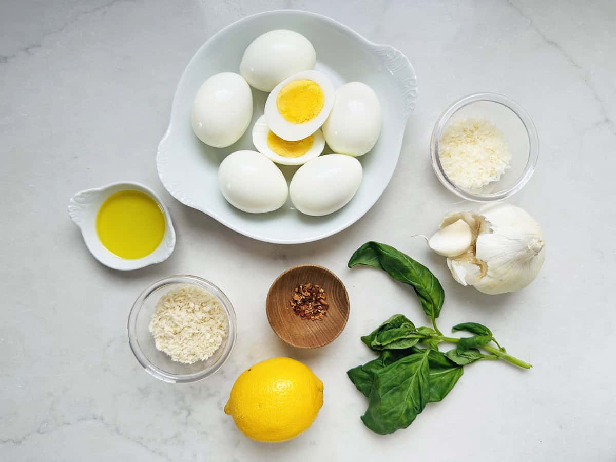 A bowl of hard-boiled eggs, a halved lemon, fresh basil leaves, a garlic bulb, grated cheese, olive oil, breadcrumbs, and red pepper flakes are arranged on a white surface.