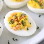 A close-up of a deviled egg topped with chopped herbs and crunchy crumbs, served on a white plate with more deviled eggs in the background.