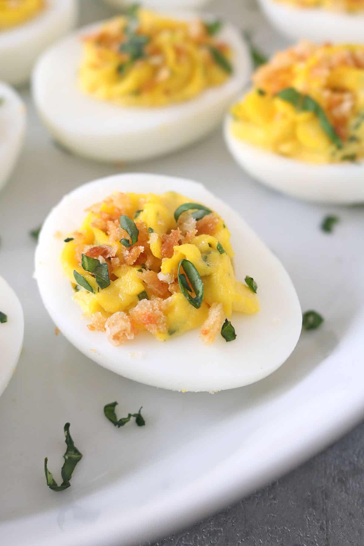 A close-up of a deviled egg topped with chopped herbs and crunchy crumbs, served on a white plate with more deviled eggs in the background.