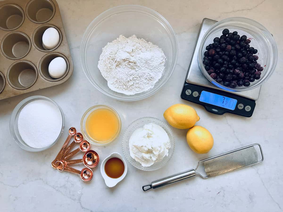 A baking setup with eggs, flour, sugar, melted butter, sour cream, vanilla, fresh lemons, a grater, blueberries on a kitchen scale, and copper measuring spoons, all arranged on a white countertop.
