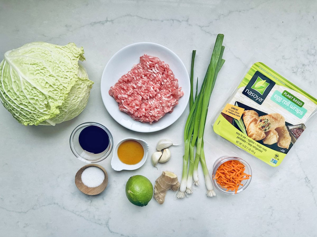 A variety of ingredients for making egg rolls are arranged on a white surface, including ground meat, a package of egg roll wraps, cabbage, green onions, grated carrots, soy sauce, garlic, ginger, a lime, salt, and oil.