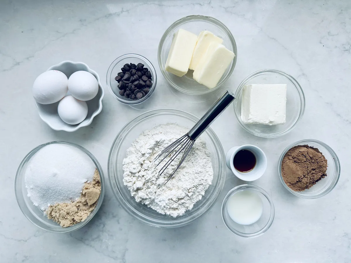 A top-down view of baking ingredients on a marble surface, including eggs, chocolate chips, butter, cream cheese, sugar, brown sugar, flour with a whisk, vanilla extract, cocoa powder, and milk in small bowls.
