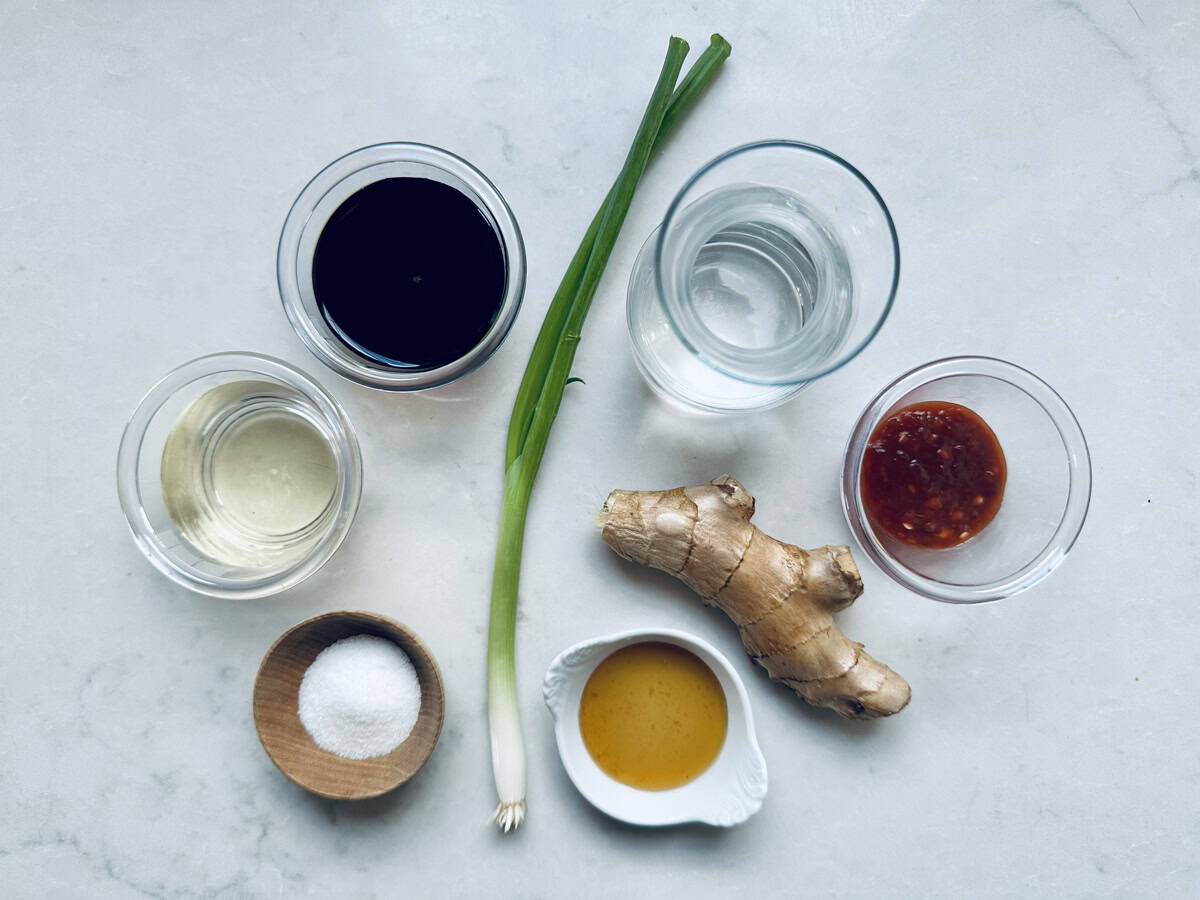 Six small bowls with soy sauce, rice vinegar, water, chili sauce, sugar, and sesame oil surround a fresh ginger root and a green onion, all arranged on a white marble surface.