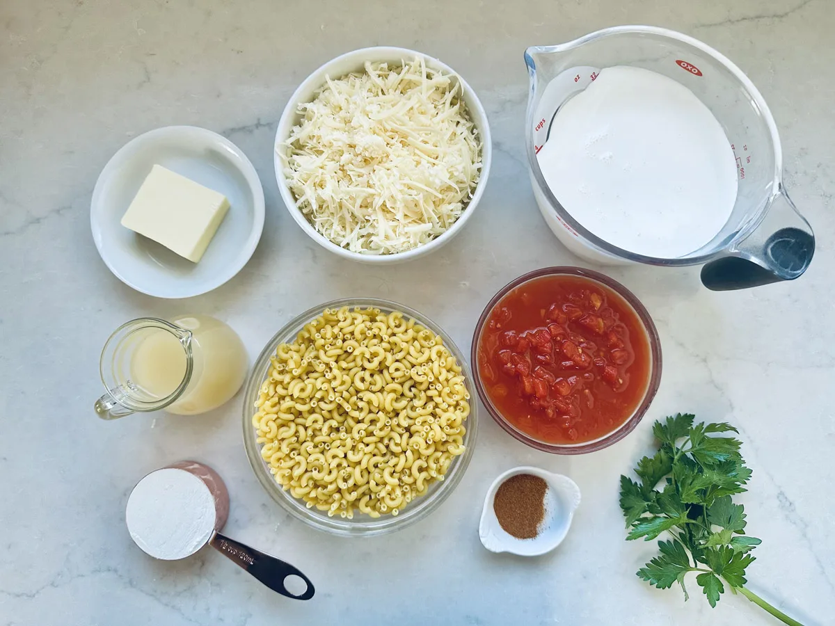 Overhead view of ingredients on a white surface: shredded cheese, milk in a glass measuring cup, diced tomatoes, elbow macaroni, chopped parsley, flour, a stick of butter, broth in a small pitcher, and a spice in a small bowl.