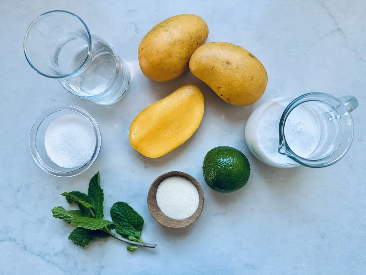Ingredients for a mango drink on a white surface: two whole mangoes, one mango half, a whole lime, a glass of water, a cup of sugar, a small bowl of powder, fresh mint leaves, and a glass jug of milk.