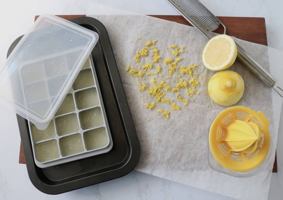 An ice cube tray filled with lemon juice sits on a tray next to grated lemon zest on parchment, a zester, a halved lemon, and a yellow citrus juicer on a wooden board.