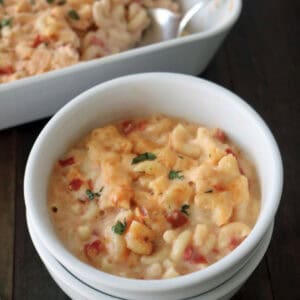 A white bowl filled with creamy macaroni and cheese topped with chopped herbs, sitting in front of a casserole dish containing more of the same macaroni and cheese on a dark wooden table.