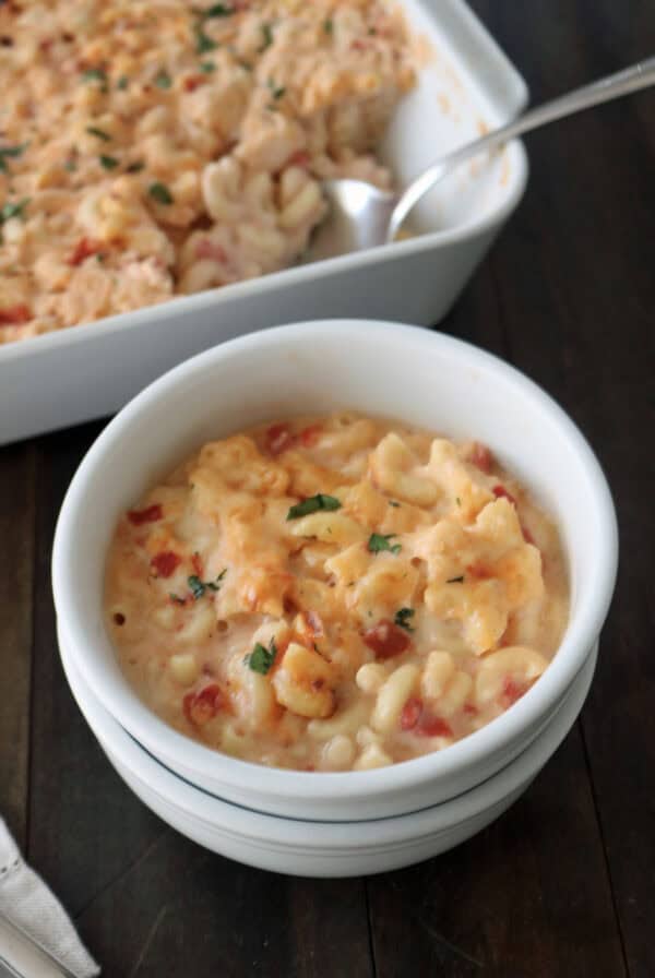 A white bowl filled with creamy macaroni and cheese topped with chopped herbs, sitting in front of a casserole dish containing more of the same macaroni and cheese on a dark wooden table.