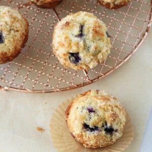 Five golden brown blueberry muffins, some resting on a round metal cooling rack and one on a crinkled paper liner, displayed on a beige surface.