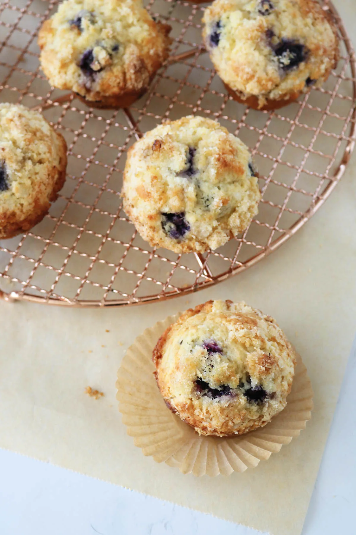 Five golden brown blueberry muffins, some resting on a round metal cooling rack and one on a crinkled paper liner, displayed on a beige surface.