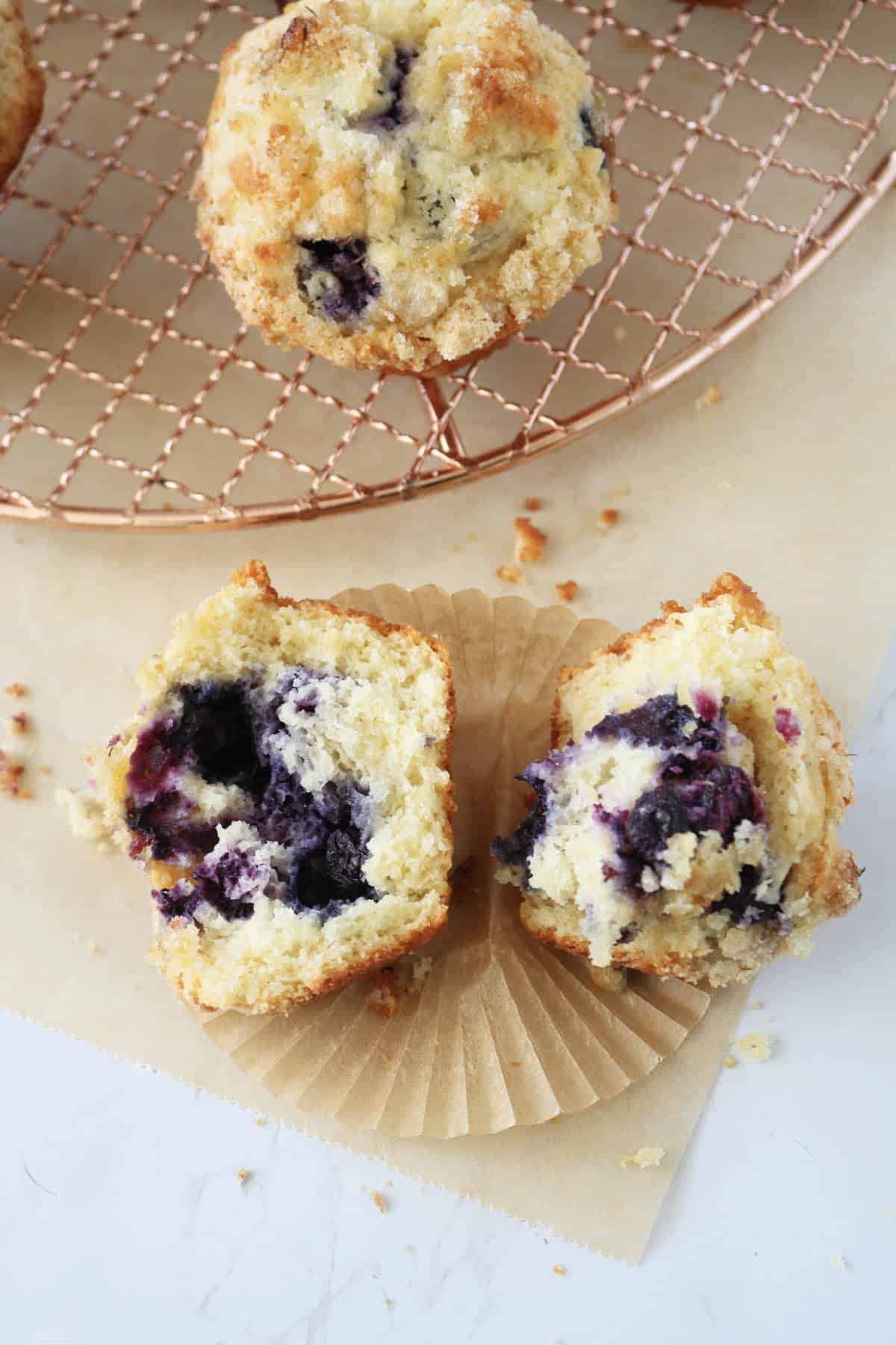 A blueberry muffin split in half sits on brown parchment paper, revealing a fluffy interior with visible blueberries. More muffins rest on a wire cooling rack in the background.