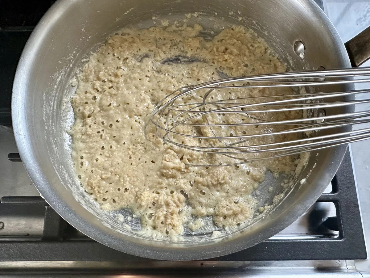 A metal whisk stirring a bubbling mixture of flour and fat in a stainless steel pan on a stovetop, making a light brown roux.