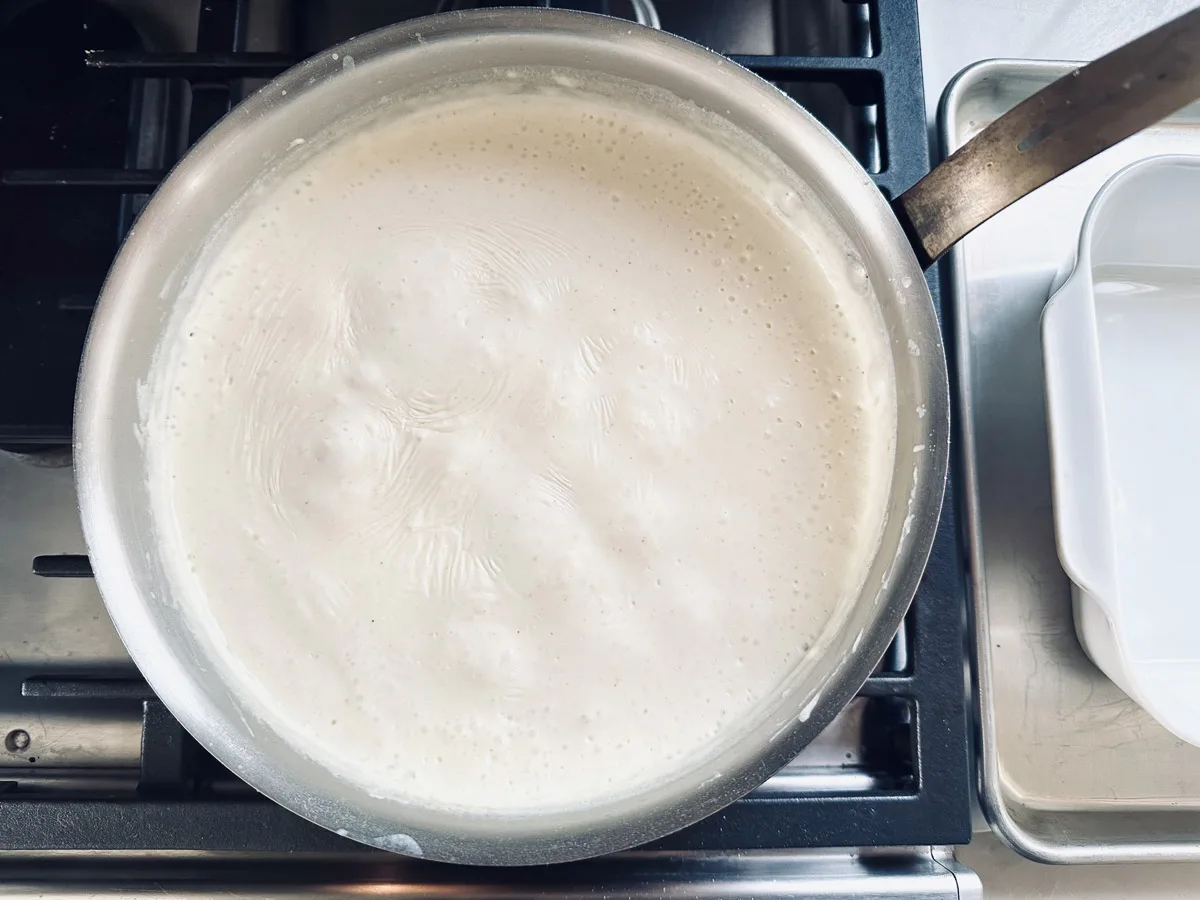 A saucepan filled with a bubbling white creamy sauce heating on a stovetop, with a metal handle visible and a white rectangular dish nearby.