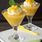 Two glass cups of mango mousse topped with whipped cream, diced mango, and mint leaves, served on a white tray with yellow and green spoons. The background is dark and the setting is elegant.