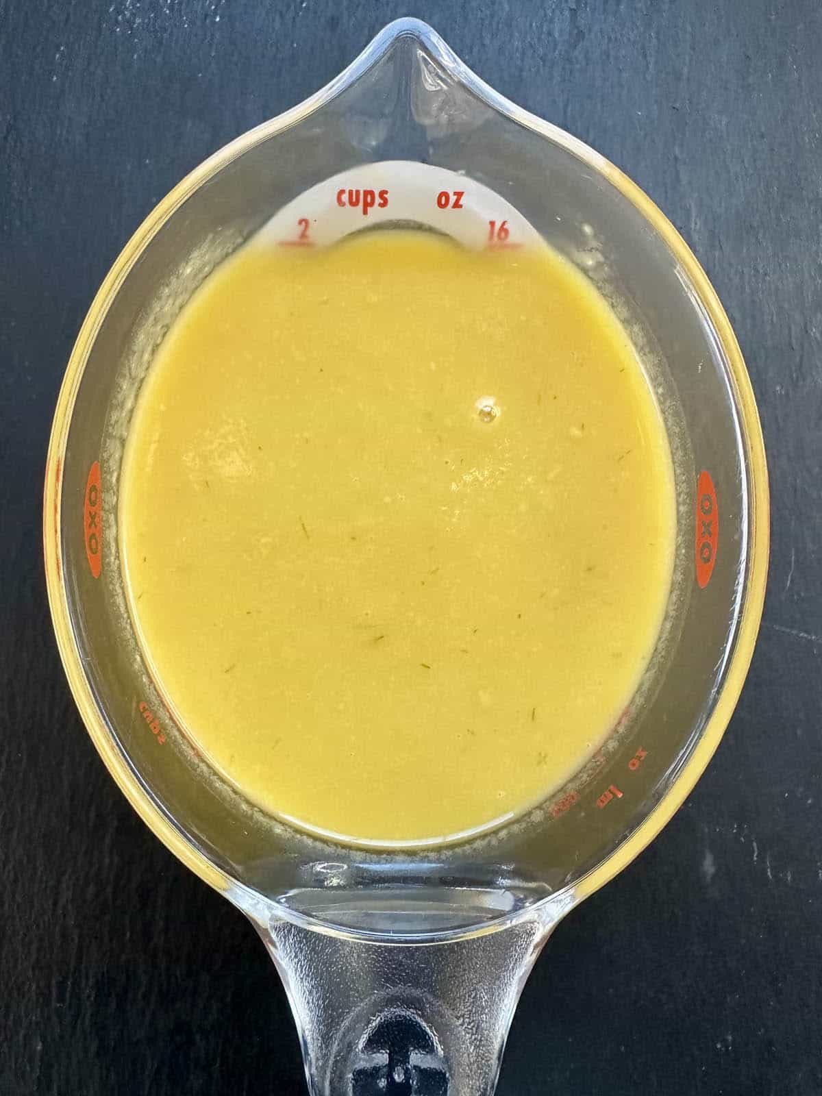 A glass measuring cup filled with a thick, yellow liquid, positioned on a dark surface. The cup shows measurements in cups and ounces along the rim.