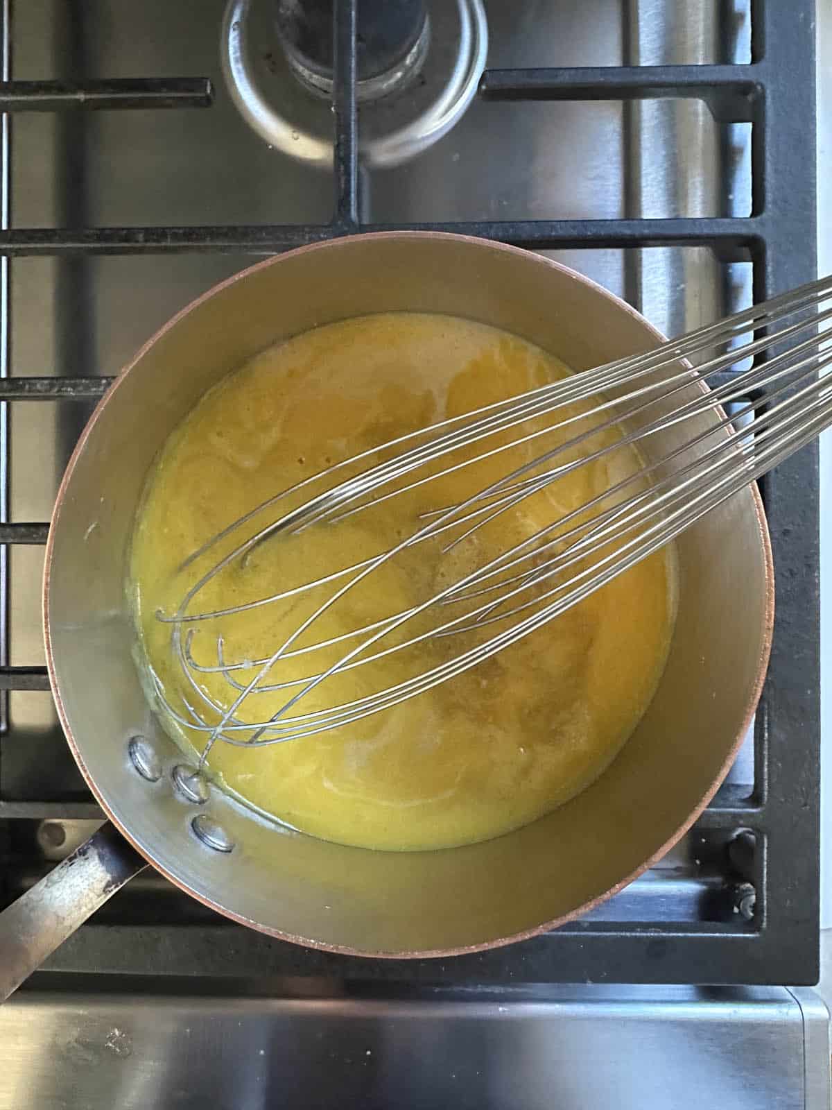 A metal whisk stirs a yellow mixture in a saucepan on a stove, viewed from above. The saucepan is positioned over a burner, and part of the stovetop grate is visible.