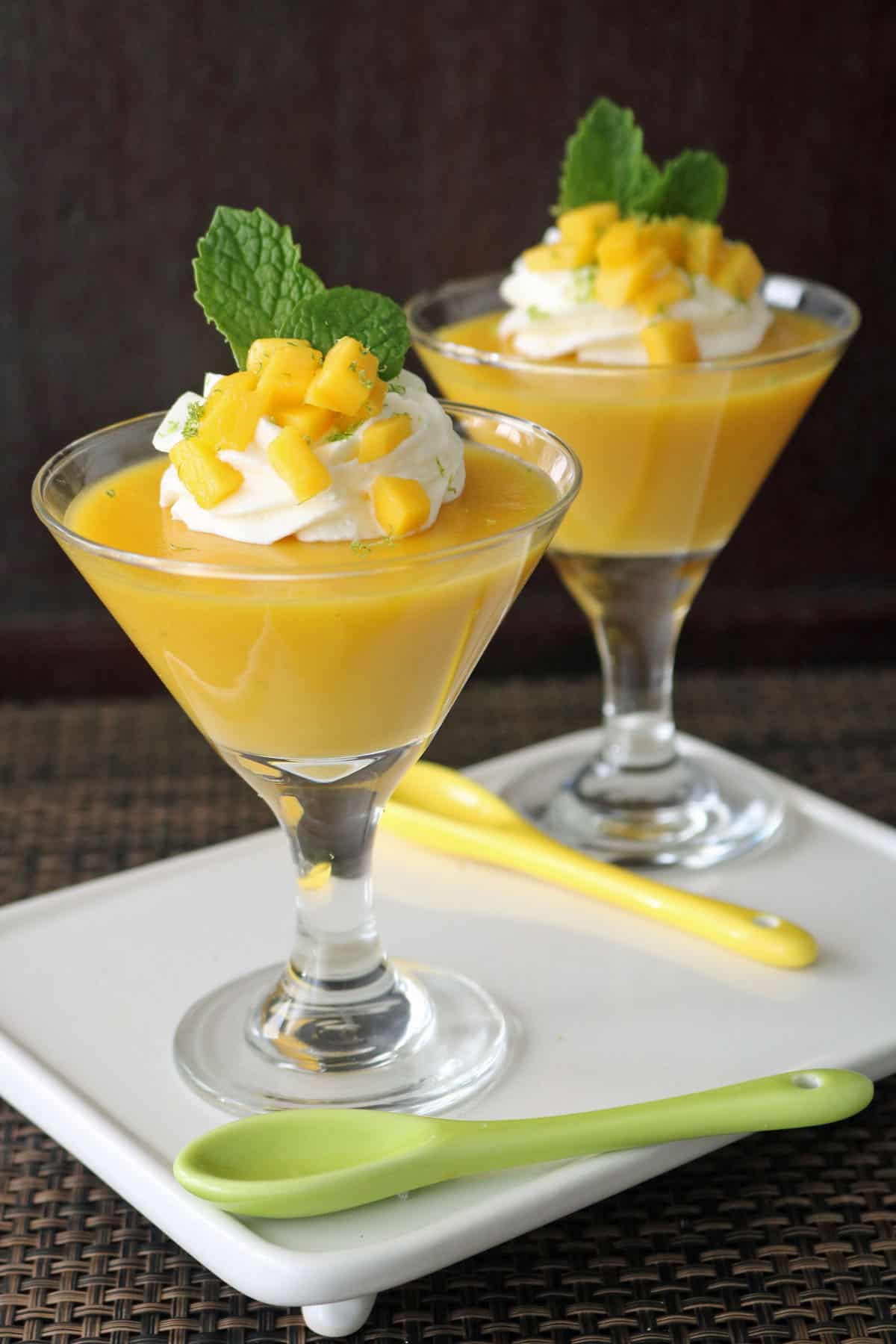 Two glass cups of mango mousse topped with whipped cream, diced mango, and mint leaves, served on a white tray with yellow and green spoons. The background is dark and the setting is elegant.