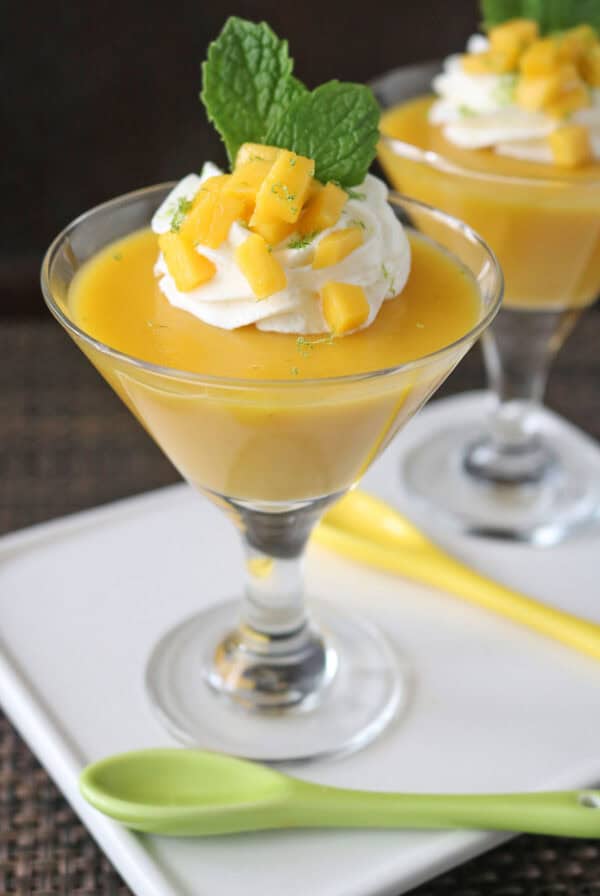 A glass dessert cup filled with mango mousse, topped with whipped cream, diced mango pieces, and a fresh mint leaf, served on a white tray with green and yellow spoons.