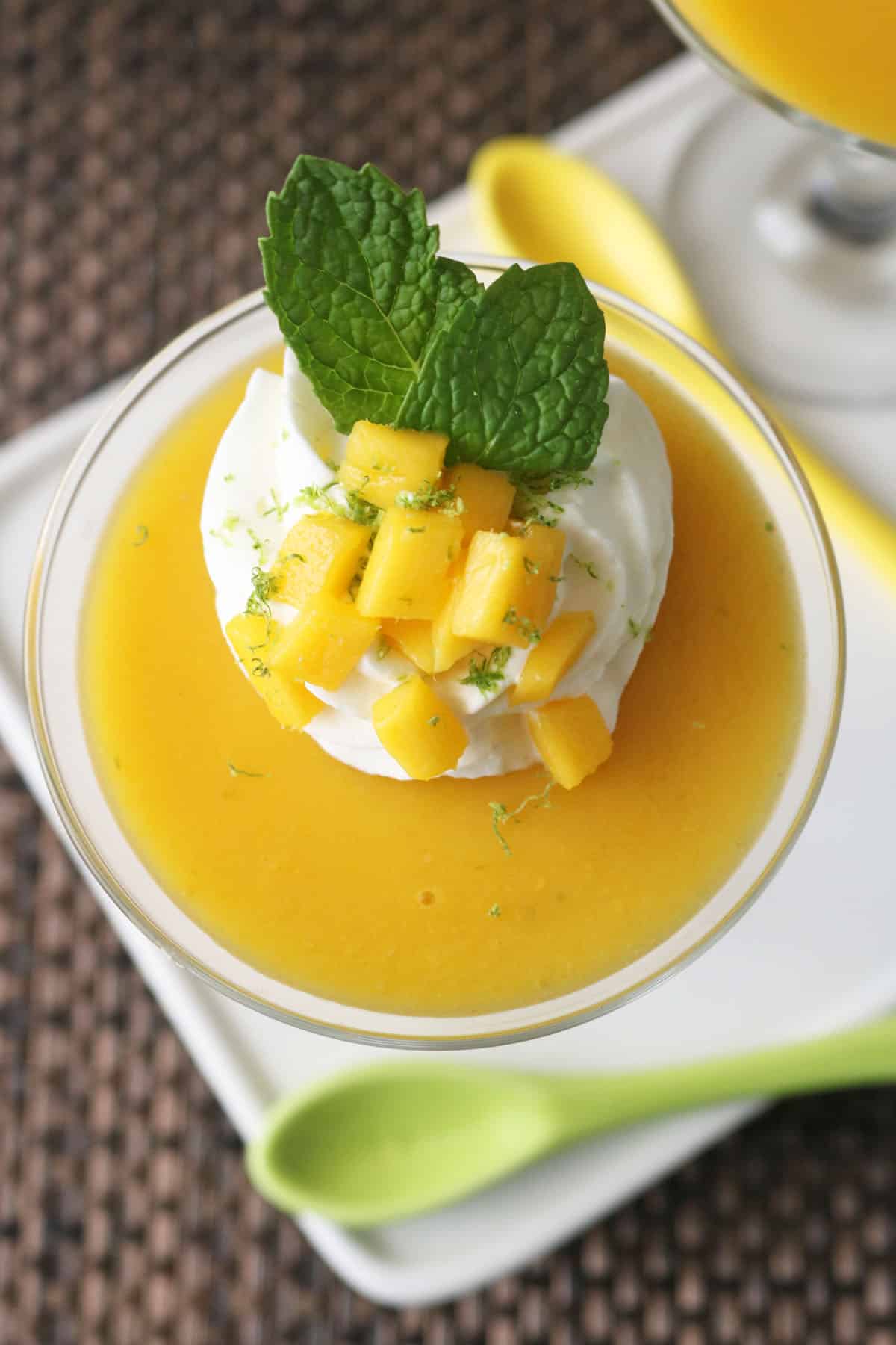 A glass dessert dish filled with smooth mango puree, topped with a swirl of whipped cream, diced mango pieces, lime zest, and two fresh mint leaves, with a green spoon beside the dish.