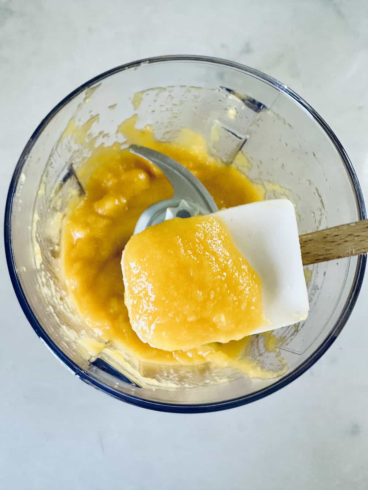 A glass blender bowl containing smooth mango puree for Mango Pudding, with a white spatula lifting some of the bright orange mixture above the bowl.