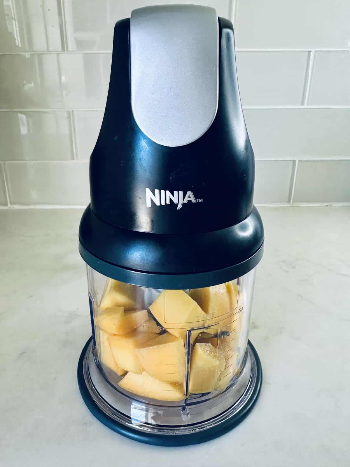 A Ninja food chopper sits on a kitchen counter, filled with chopped yellow apple pieces inside the clear container—perfect for preparing ingredients for mango pudding. White tile backsplash is visible in the background.