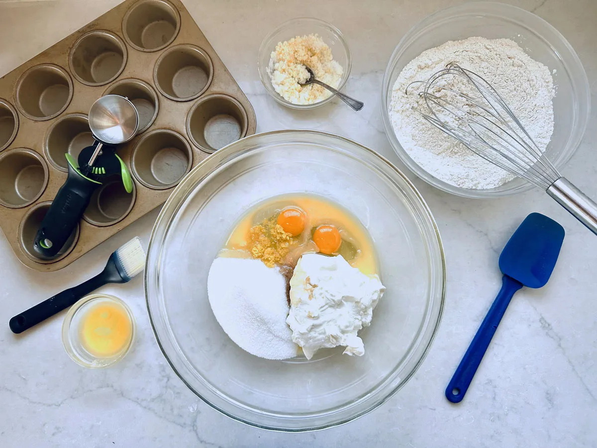 A muffin tin, ice cream scoop, brush, bowl of flour, bowl with eggs, sugar, and sour cream, small bowls of cheese and egg wash, and a spatula are arranged on a white countertop for baking.