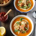 Two bowls of orange soup topped with croutons, cilantro, and a swirl of cream, placed on a wooden table with a jar of red sauce, lemon wedges, a bowl of croutons, and spoons nearby.