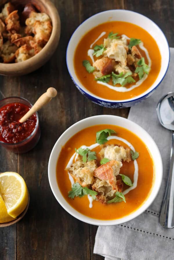 Two bowls of orange soup topped with croutons, cilantro, and a swirl of cream, placed on a wooden table with a jar of red sauce, lemon wedges, a bowl of croutons, and spoons nearby.