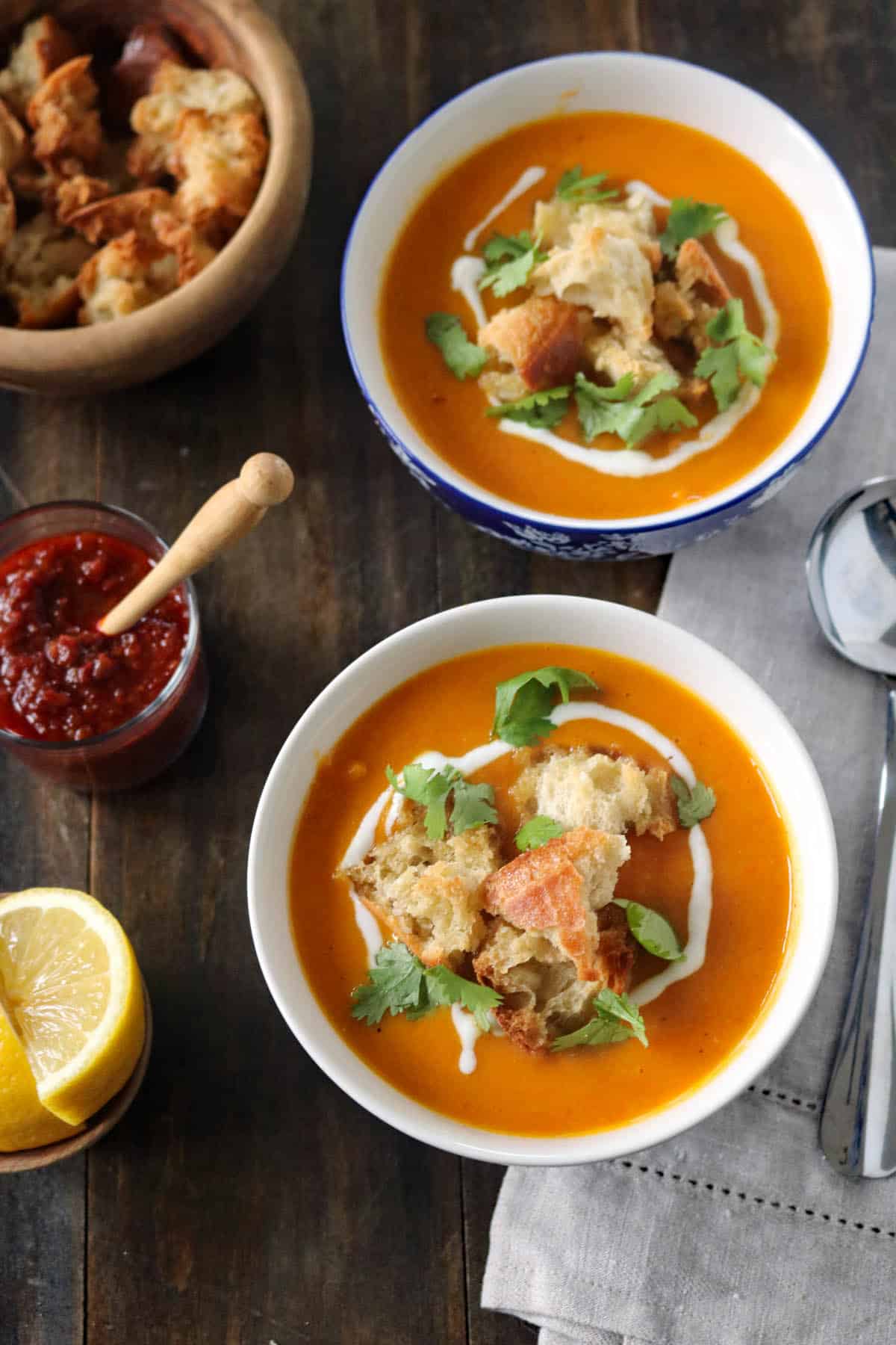 Two bowls of orange soup topped with croutons, cilantro, and a swirl of cream, placed on a wooden table with a jar of red sauce, lemon wedges, a bowl of croutons, and spoons nearby.