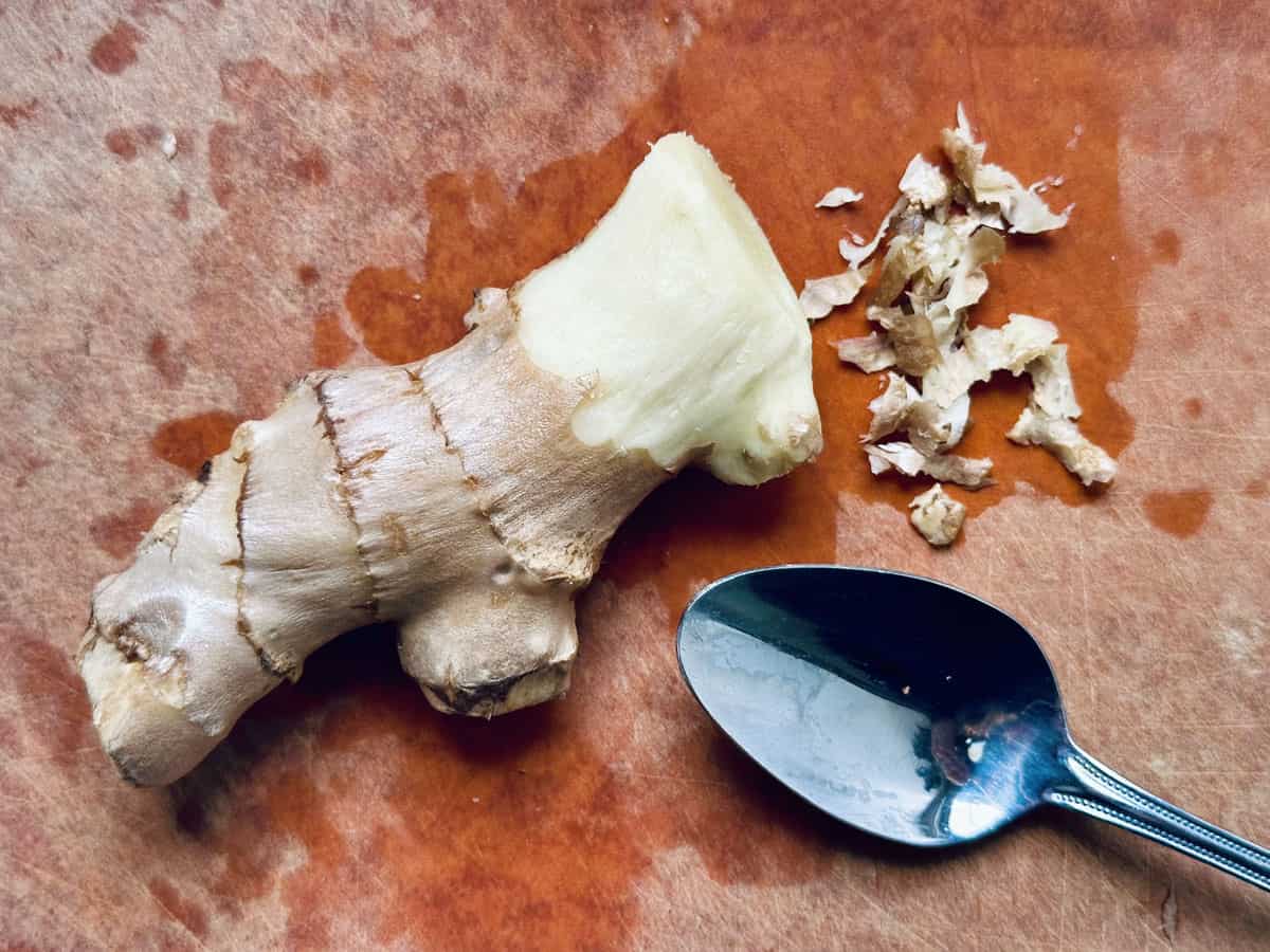 A piece of fresh ginger root with some peeled skin beside it, and a metal spoon on a wooden cutting board.