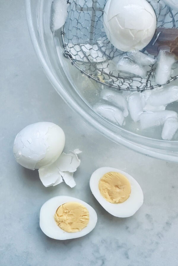 A peeled hard-boiled egg, an unpeeled egg, and a halved hard-boiled egg rest on a countertop beside a glass bowl filled with ice water and a wire strainer holding another egg.