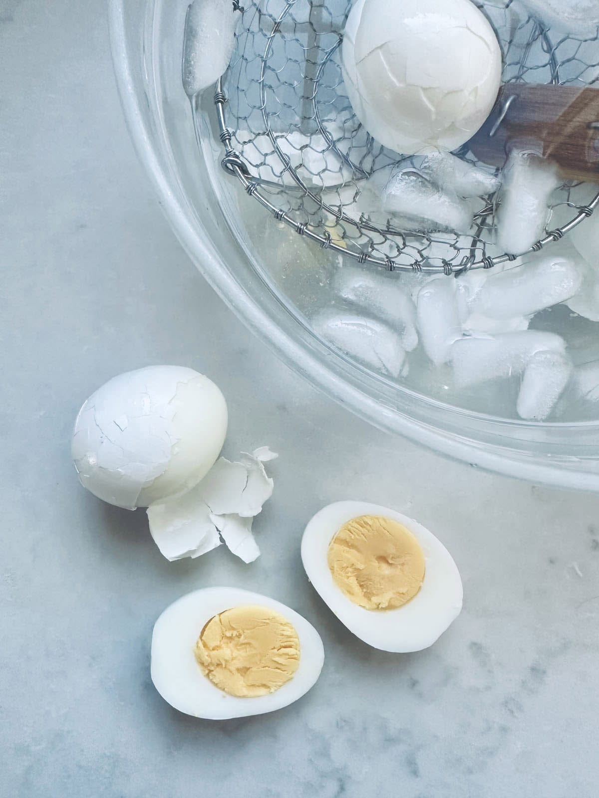 A peeled hard-boiled egg, an unpeeled egg, and a halved hard-boiled egg rest on a countertop beside a glass bowl filled with ice water and a wire strainer holding another egg.