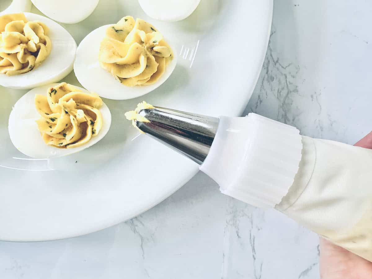 A hand pipes yellow filling into halved hard-boiled eggs on a white plate, making deviled eggs. The scene is set on a white marble surface.