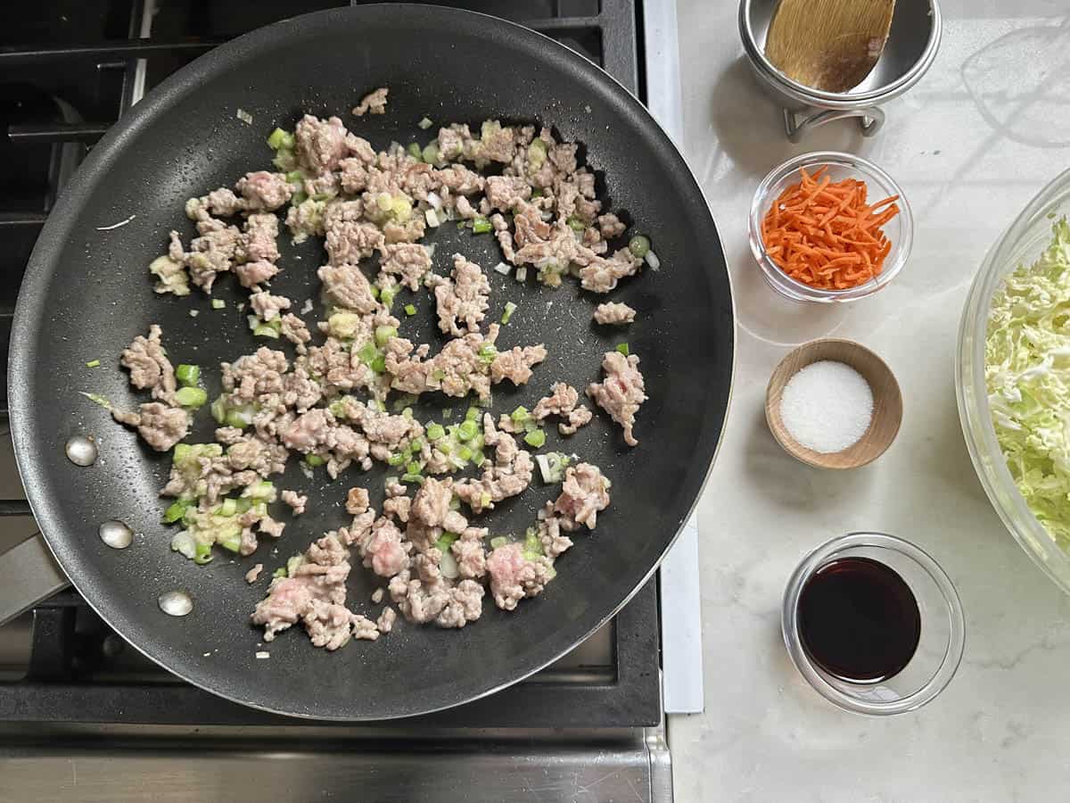 A skillet with ground meat and chopped green vegetables cooking on a stove. Nearby are small bowls containing shredded carrots, salt, soy sauce, and a large bowl of shredded cabbage on a white countertop.
