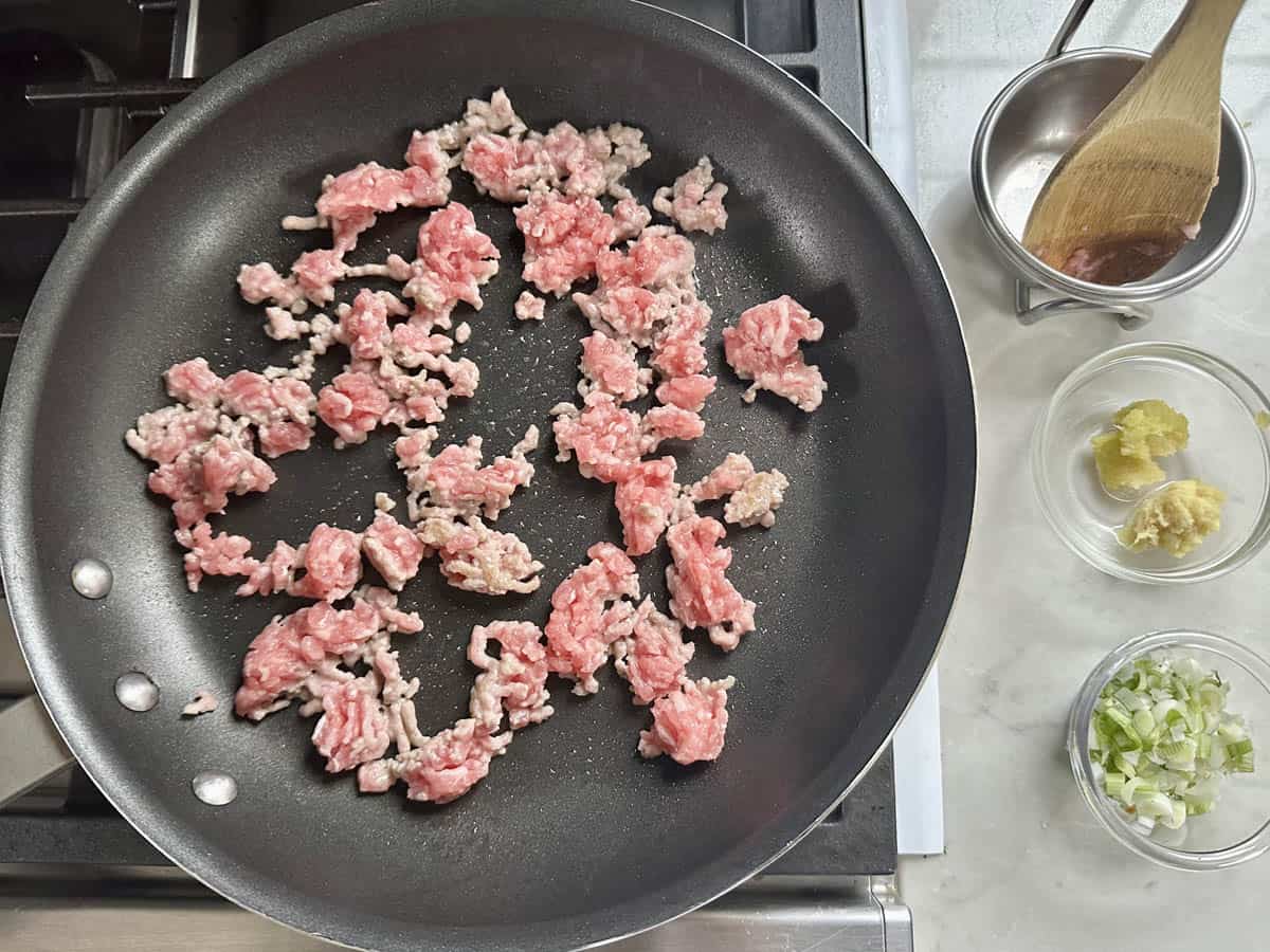 Ground meat is being cooked in a large nonstick skillet on a stove. Beside the skillet, there are small bowls with chopped green onions, minced garlic, and grated ginger, along with a wooden spoon in a metal bowl.