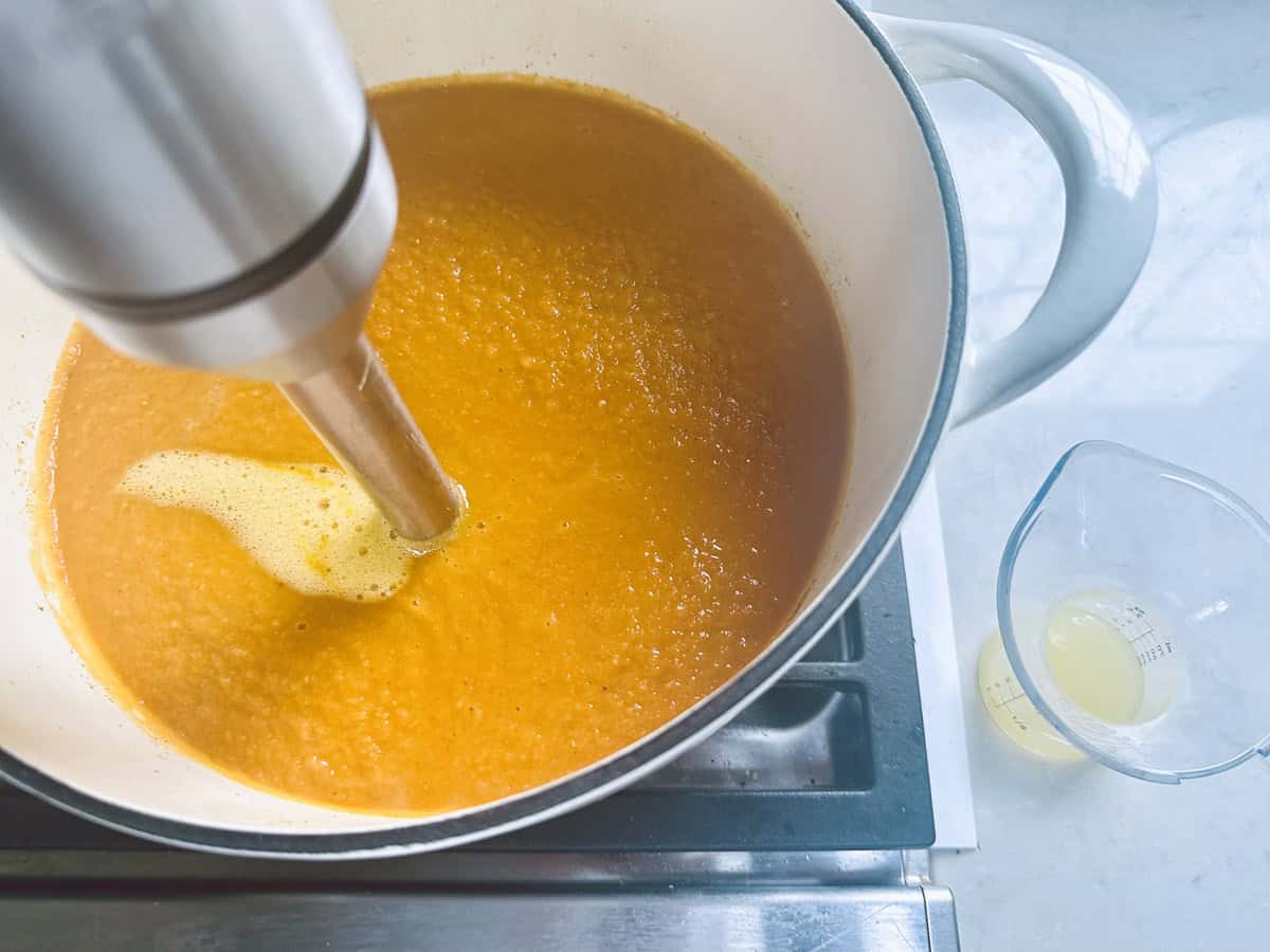 A hand blender is pureeing orange soup in a white pot on a stove. Next to the pot is a small measuring cup containing yellow liquid.