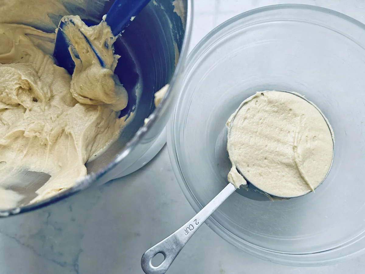 A metal mixing bowl with cookie dough and a blue spatula beside a clear bowl containing a scoop of dough on a white surface. A metal measuring cup, labeled 2 cup, holds the dough in the clear bowl.