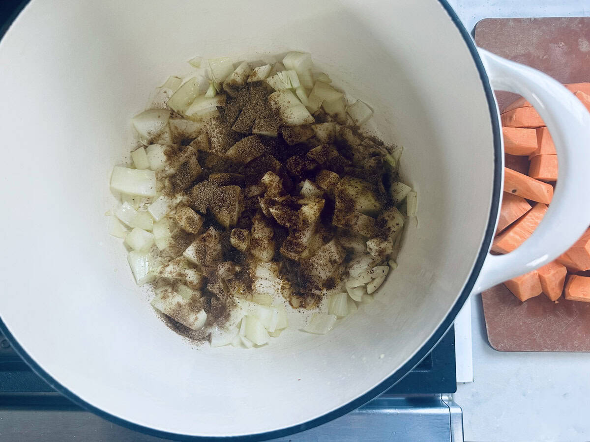 Chopped onions with spices are cooking in a large white pot on a stove; chopped sweet potatoes are on a brown cutting board nearby.
