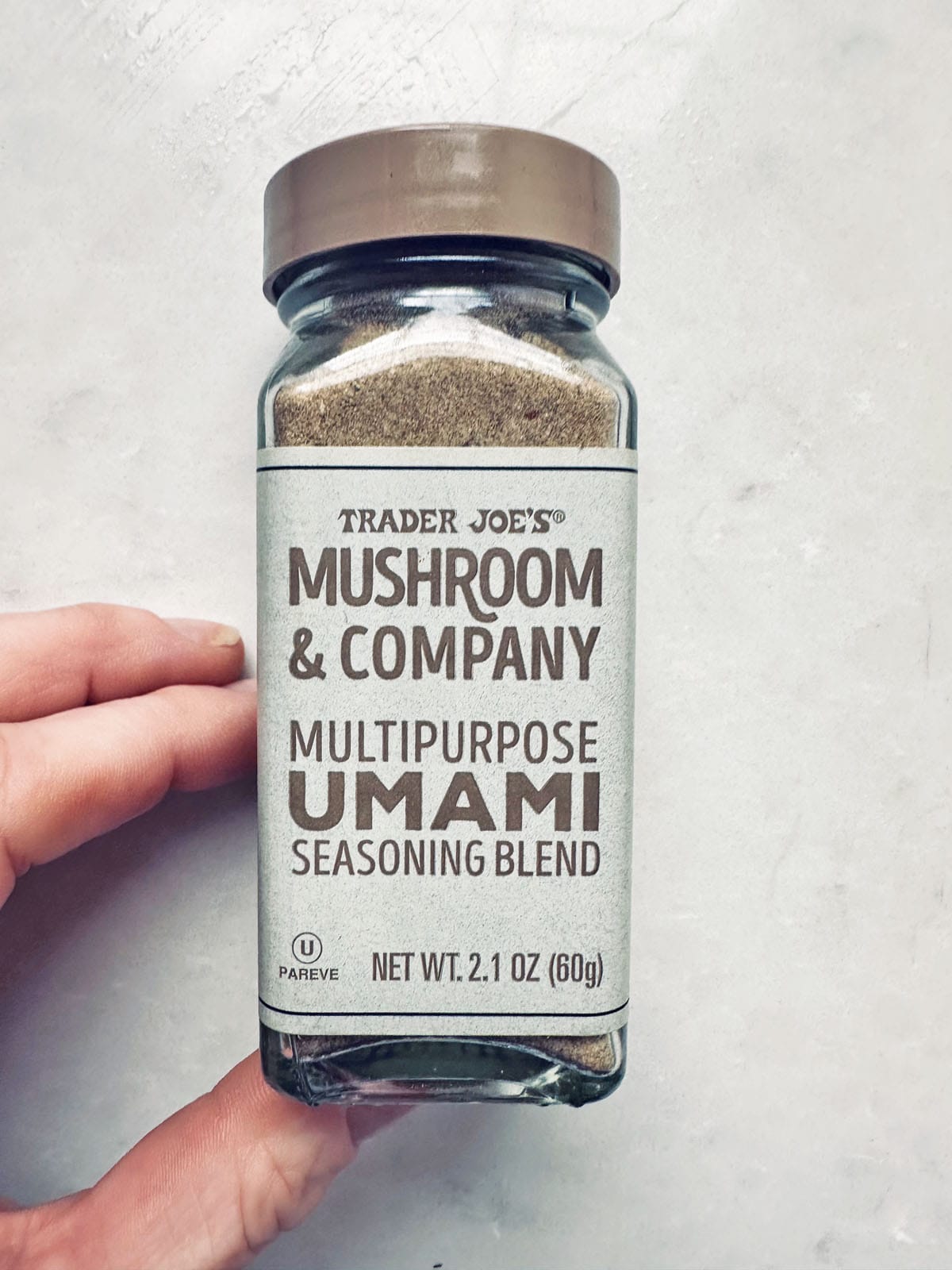 A hand holds a glass jar of Trader Joe’s Mushroom & Company Multipurpose Umami Seasoning Blend against a light background. The label is beige with brown text and a brown lid tops the jar.