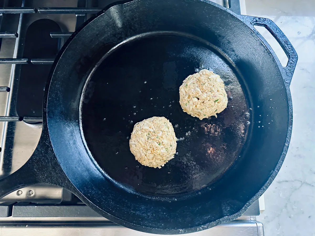 Two uncooked crab cakes are placed in a black cast iron skillet on a stovetop, ready to be cooked. The skillet sits on a gas burner, and the kitchen counter is visible to the side.