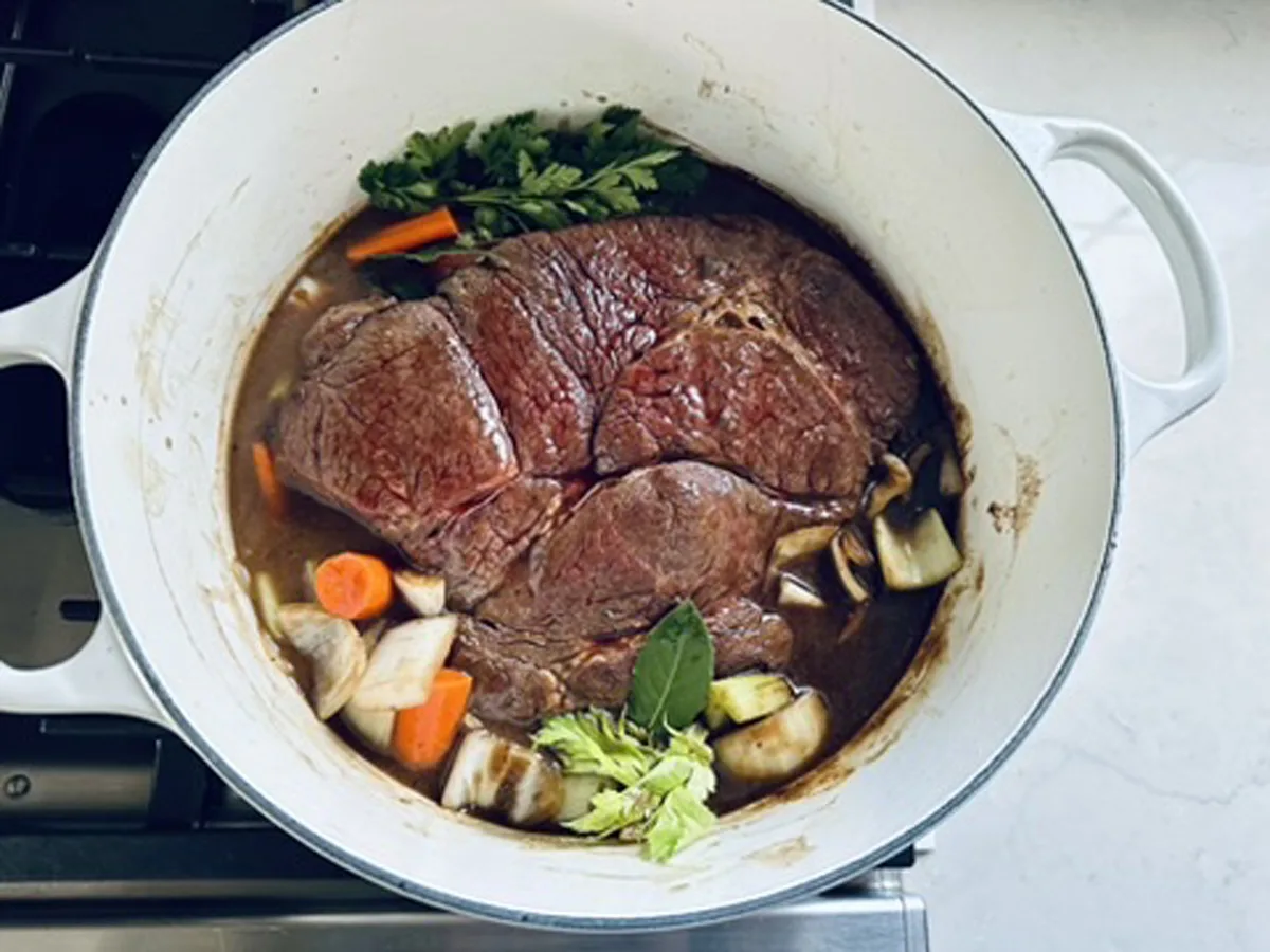 A large piece of beef is searing in a white Dutch oven with chopped carrots, celery, onions, fresh herbs, and bay leaves, surrounded by brown cooking liquid on a stovetop.