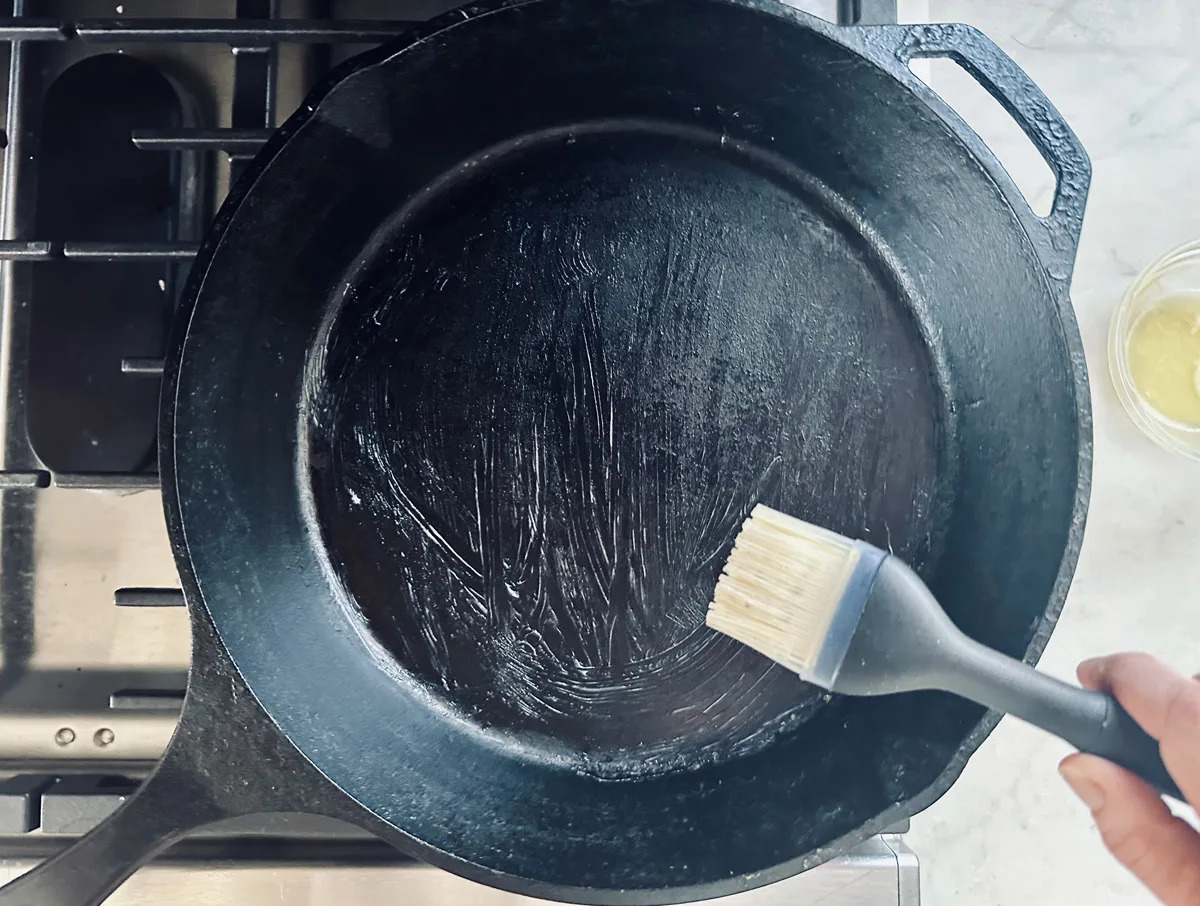 A hand uses a silicone brush to spread oil or butter onto a black cast iron skillet on a stovetop. A small bowl with liquid is visible nearby on a light-colored countertop.