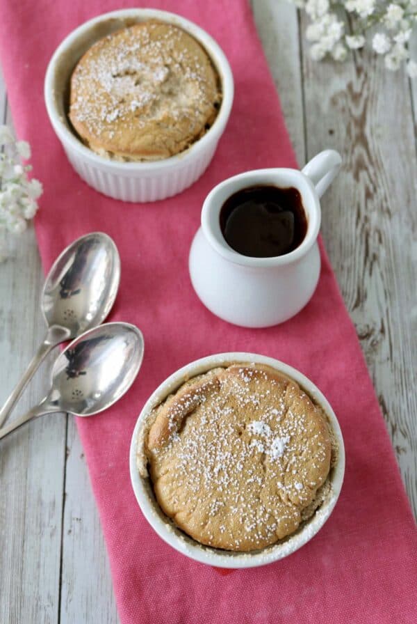 Two small ramekins with golden, powdered sugar-dusted desserts on a pink napkin, accompanied by two silver spoons and a small white pitcher of chocolate sauce on a rustic wooden surface. White flowers decorate the corners.