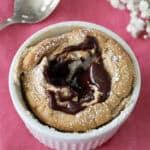 A chocolate lava cake in a white ramekin, topped with melted chocolate and powdered sugar, sits on a pink cloth next to a silver spoon and white flowers.