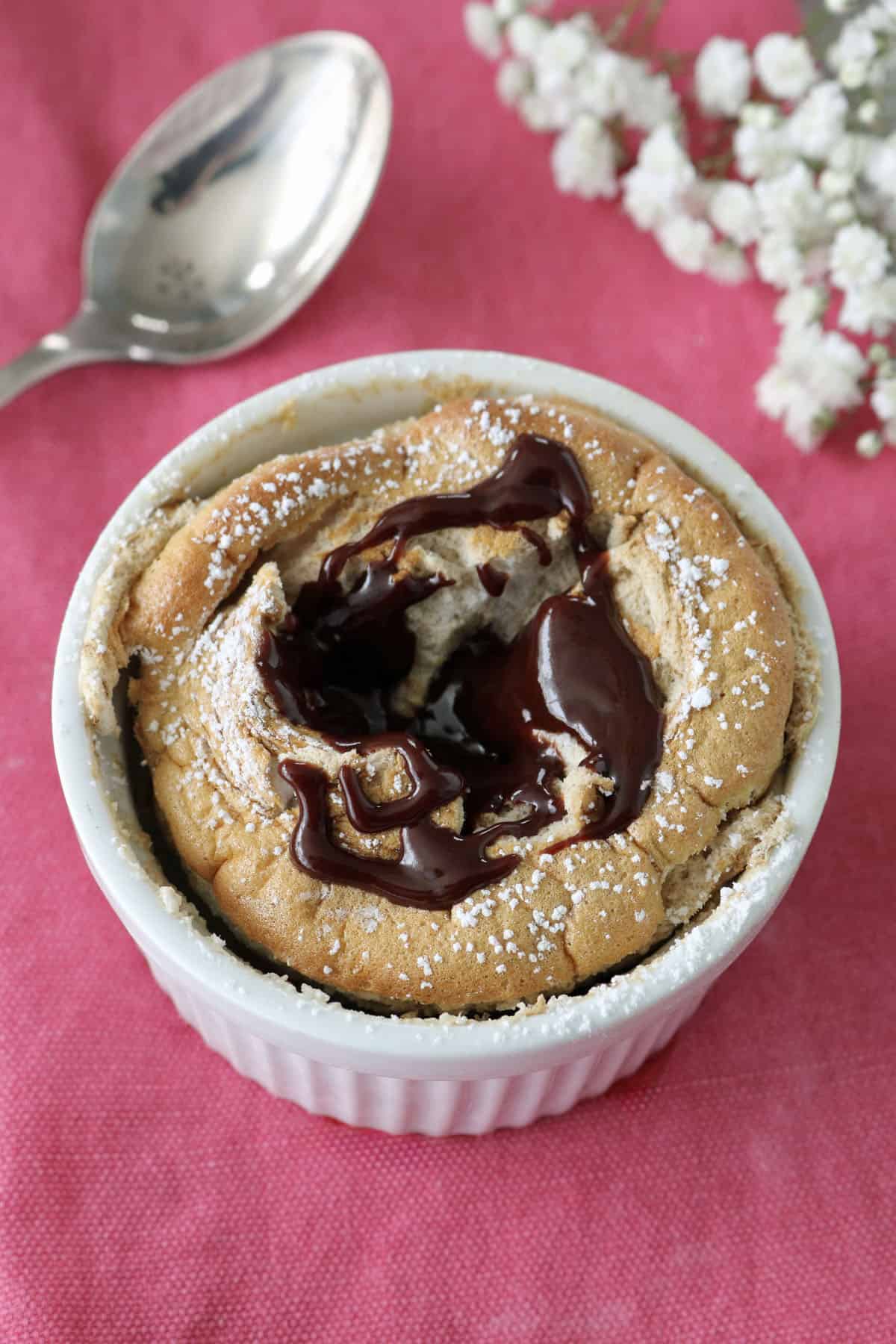 A chocolate lava cake in a white ramekin, topped with melted chocolate and powdered sugar, sits on a pink cloth next to a silver spoon and white flowers.