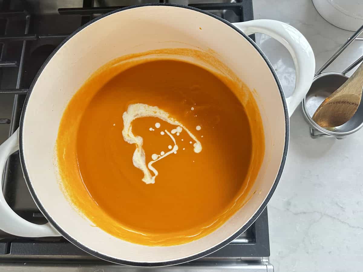 A white pot on a stovetop contains creamy orange soup, likely tomato or carrot, with a swirl of cream on top. A wooden spoon and utensil holder are visible on the counter beside the pot.